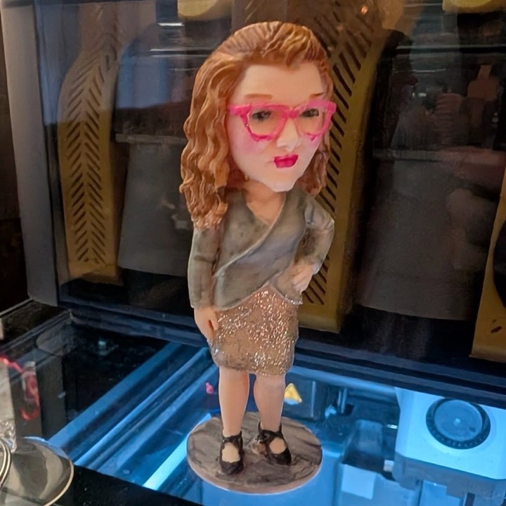 Full-color 3D printed figure of a woman with curly hair and pink glasses standing on a round base