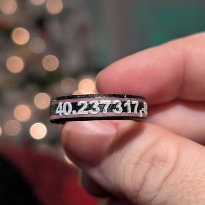3D printed black ring with GPS coordinates in white lettering, held between two fingers