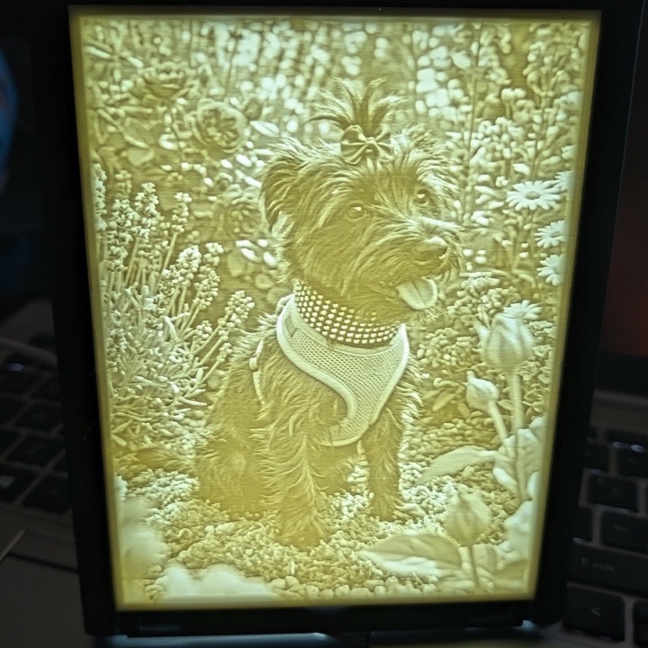 Backlit 3D printed lithophane of a small dog sitting in a garden, glowing warm yellow in a black frame