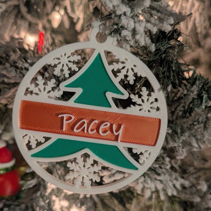3D printed round Christmas ornament with a green tree, snowflakes and a name banner